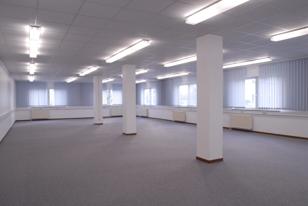 Geräumiger Büroraum mit hellen Fenstern und grauem Teppichboden, ausgestattet mit Deckenleuchten.